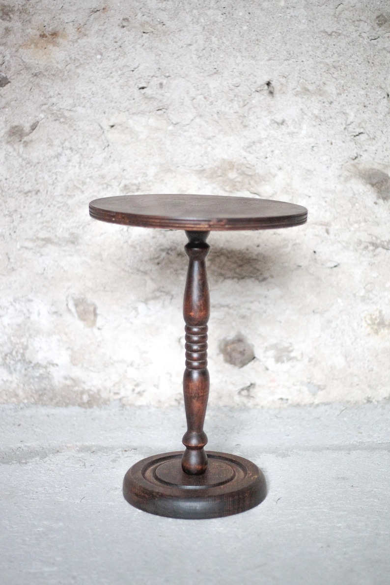 Pode incluir: Uma mesa de pedestal de madeira castanho-escura com um tampo circular, uma coluna central torneada com sulcos decorativos e uma base redonda e plana. O veio da madeira &eacute; vis&iacute;vel em toda a estrutura, exibindo um acabamento tingido profundo.