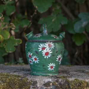 May include: A green ceramic water jug with a handle and two spouts. The jug is decorated with white daisies with red centers. The background is blurred green foliage.