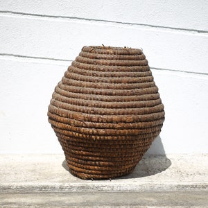 May include: A large, brown, woven basket made of natural materials. The basket is round and has a wide opening at the top. It is sitting on a concrete surface.