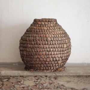 May include: A round, woven basket with a narrow opening at the top. The structure is composed of thick, dark brown natural fibers coiled and bound together with thinner, darker cordage, creating a textured, layered appearance throughout the body.