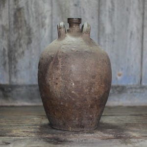 Pode incluir: Um jarro rústico de cerâmica castanha com corpo arredondado e duas pequenas pegas. O jarro tem um gargalo estreito e um aspeto escuro e envelhecido. O fundo é uma superfície de madeira desgastada.