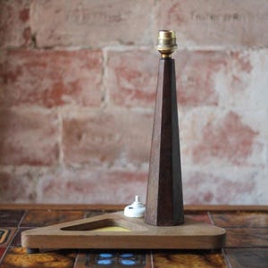 Könnte beinhalten: Ein Tischlampenfuß mit einer hohen, dunkelbraunen, konisch zulaufenden Holzsäule und einer Metallfassung an der Spitze. Die Säule ist auf einem dreieckigen Holzsockel montiert, der einen kleinen weißen Schalter nahe der Säulenbasis aufweist.