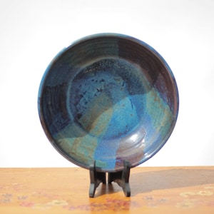 Op de afbeelding: Een ronde keramische kom met een glanzend glazuur in gemengde tinten diep marineblauw, koningsblauw en aards bruin. Het binnenoppervlak heeft een gladde, reflecterende afwerking met subtiele kleurovergangen over de gehele ronde kom.