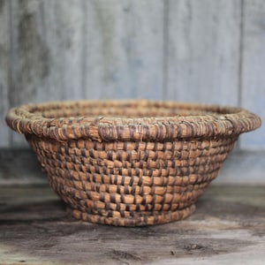 May include: A round, woven basket with a wide, shallow bowl shape. The basket is made of natural-colored fibers, with a textured, open weave. The rim is thicker and more defined. The basket sits on a wooden surface.