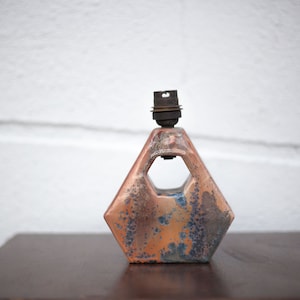 Puede incluir: Una lámpara de mesa de cerámica con una base geométrica hexagonal. La base es una mezcla moteada de azul, naranja y marrón. La lámpara tiene un simple casquillo de metal negro.