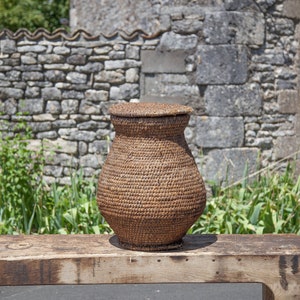 Puede incluir: Una cesta de mimbre tejida con tapa, sentada sobre una superficie de madera. La cesta es marrón y tiene un aspecto natural y rústico. Está delante de una pared de piedra y vegetación verde.