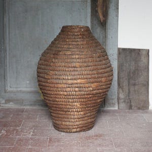 May include: A large, woven basket with a bulbous shape and a narrow neck. The basket is made of natural-colored fibers, with horizontal bands creating a textured surface. It stands on a red brick floor against a weathered gray door.