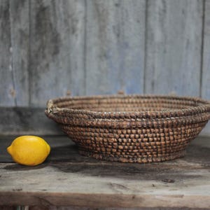 May include: A woven brown basket with a rounded shape sits on a wooden surface. A bright yellow lemon rests beside the basket. The background is a weathered, blue-toned wooden wall.