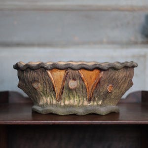 Peut inclure: Un pot rectangulaire rustique avec une texture imitant l'écorce. Il présente un bord ondulé et trois motifs triangulaires de couleur orange. La palette de couleurs comprend des bruns, des verts et des oranges, évoquant un thème naturel et terreux.