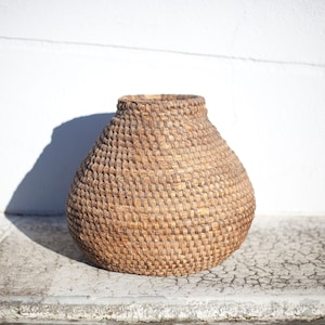 May include: A large, round, brown woven basket made of natural materials. The basket has a wide opening and a rounded bottom. It is sitting on a cracked, light brown surface.
