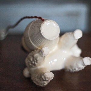 May include: A vintage ceramic poodle figurine, lying on its back, with a cream-colored body and a textured, cylindrical basket on its back. The poodle's paws are visible, and a thin, dark wire extends from the basket.