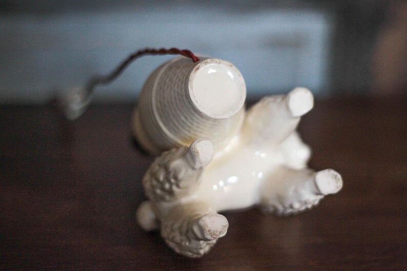 May include: A vintage ceramic poodle figurine, lying on its back, with a cream-colored body and a textured, cylindrical basket on its back. The poodle's paws are visible, and a thin, dark wire extends from the basket.