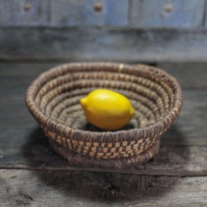 May include: A woven basket with a yellow lemon inside. The basket is made of natural fibers in shades of brown and tan, with a checkered pattern on the base. The lemon is bright yellow.