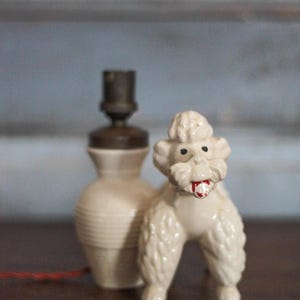 May include: A vintage ceramic poodle figurine with a red tongue stands next to a cream-colored lamp. The lamp has a brown top and a ribbed base. The poodle is white with black eyes.