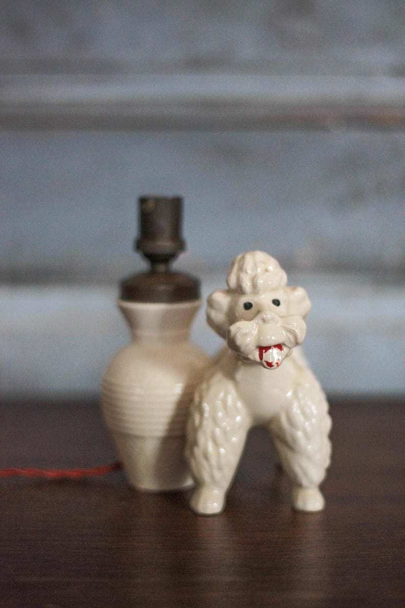 May include: A vintage ceramic poodle figurine with a red tongue stands next to a cream-colored lamp. The lamp has a brown top and a ribbed base. The poodle is white with black eyes.