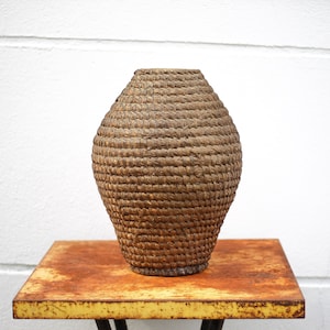 May include: A woven brown vase with a rounded shape sits on a rusty metal table.