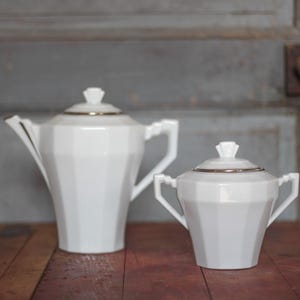 Peut inclure: Service à thé en porcelaine blanche au design géométrique. L'ensemble comprend une théière et un sucrier, tous deux avec des bordures dorées. La théière est de forme haute et élancée, tandis que le sucrier est plus petit avec deux anses.
