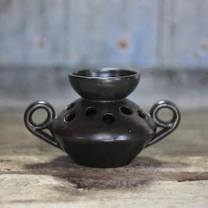 May include: A glossy black ceramic flower frog vase featuring a rounded body with multiple circular openings on the top surface, a central raised bowl-shaped opening, and two curved loop handles extending from the sides of the vessel.