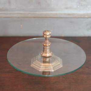 May include: A clear glass cake stand with a decorative brass-coloured metal base and stem. The round glass plate sits atop a hexagonal base and a detailed stem. The stand is displayed on a dark wooden surface.