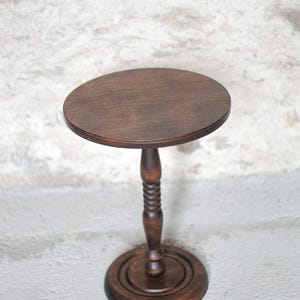 Pode incluir: Um suporte de pedestal em madeira castanho-escura com um topo circular, uma coluna central com um design torneado e canelado, e uma base redonda escalonada. A pe&ccedil;a &eacute; feita de madeira com gr&atilde;o vis&iacute;vel e um acabamento polido.