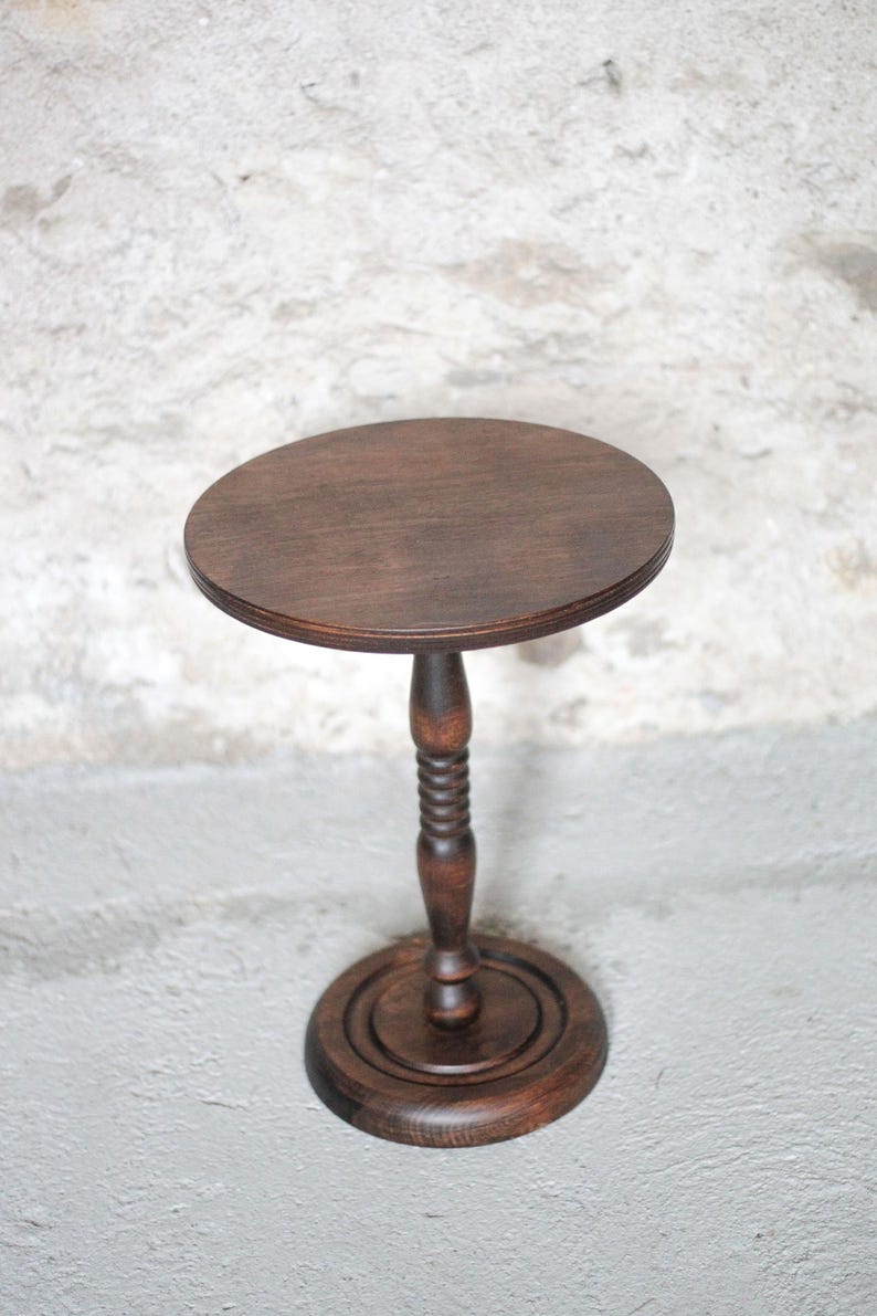 Pode incluir: Um suporte de pedestal em madeira castanho-escura com um topo circular, uma coluna central com um design torneado e canelado, e uma base redonda escalonada. A pe&ccedil;a &eacute; feita de madeira com gr&atilde;o vis&iacute;vel e um acabamento polido.