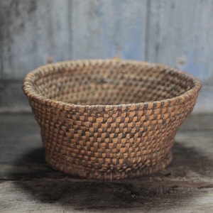 May include: A woven basket with a rounded shape and a wide opening. The basket is made of natural-colored materials, with a checkered pattern. The basket sits on a wooden surface.