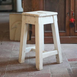 Può includere: Un piccolo sgabello in legno bianco con un sedile quadrato e quattro gambe angolate. Lo sgabello ha un aspetto invecchiato e usurato, suggerendo che sia vintage o antico. Lo sfondo comprende un armadio in legno e un pavimento piastrellato.