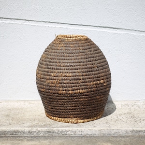 May include: A large, round, brown woven basket made of natural materials. The basket is sitting on a light gray concrete surface.