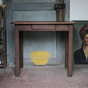 Peut inclure: Un bureau en bois marron avec un seul tiroir et un bouton rond. Le bureau est placé devant une porte grise vieillie et un portrait encadré d'une femme. Le sol est carrelé de briques rouges.