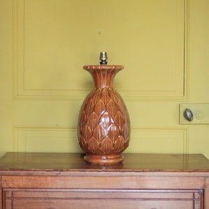 Puede incluir: Una lámpara de mesa de cerámica marrón con diseño de piña. La lámpara tiene una superficie texturizada y un casquillo de color latón. Se encuentra sobre una mesa de madera contra una pared amarilla.