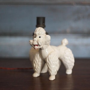 May include: A vintage white ceramic poodle figurine with a red mouth and black eyes. The poodle has a classic, stylized design with fluffy fur and is positioned on a dark wooden surface. A black lamp base is behind the poodle.