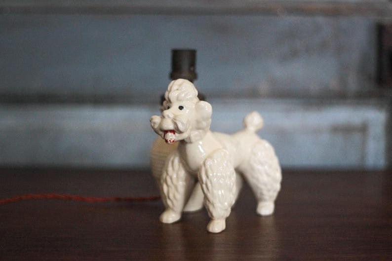 May include: A vintage white ceramic poodle figurine with a red mouth and black eyes. The poodle has a classic, stylized design with fluffy fur and is positioned on a dark wooden surface. A black lamp base is behind the poodle.