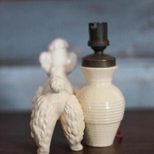May include: A vintage-style cream-colored ceramic poodle figurine stands next to a small lamp base. The lamp base has a textured, ribbed design and a dark metal top. The poodle figurine is detailed with textured fur.