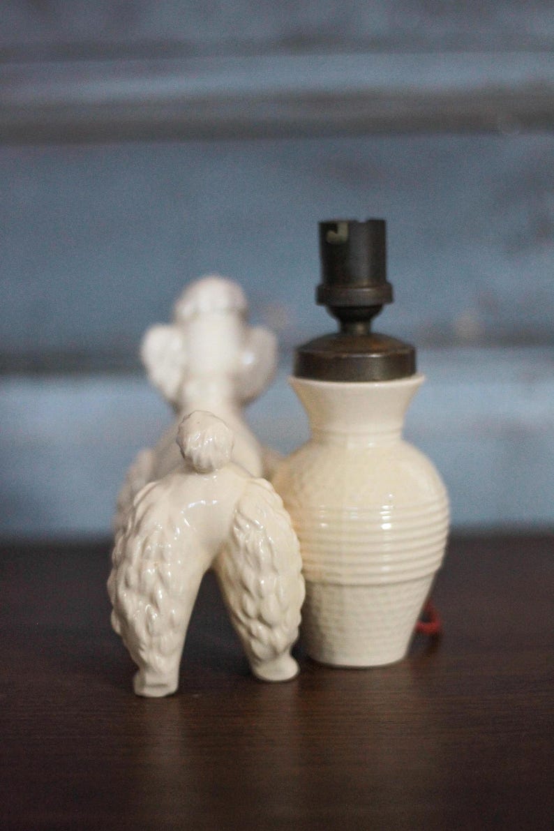 May include: A vintage-style cream-colored ceramic poodle figurine stands next to a small lamp base. The lamp base has a textured, ribbed design and a dark metal top. The poodle figurine is detailed with textured fur.