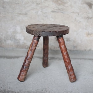 Può includere: Uno sgabello piccolo a tre gambe realizzato in legno marrone scuro. La seduta circolare presenta una superficie ruvida e irregolare con venature del legno visibili. Le tre gambe hanno la forma di rami spessi e nodosi per una base solida.