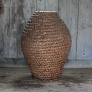 May include: A woven basket with a rounded body and a narrow opening. The basket is made of a natural fiber, with a textured surface and a warm brown color. The basket is displayed on a wooden surface.