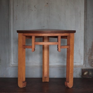 May include: A small, three-legged wooden table with a triangular top. The top is dark brown, while the legs and supports are a lighter wood tone. The table has a minimalist design, suitable for a small space or as a decorative accent piece.