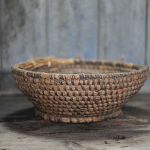 May include: A handcrafted, round basket with a wide, shallow bowl shape. The basket is woven from natural fibers in shades of brown and tan, with a textured, patterned surface. The basket sits on a small, rounded base.