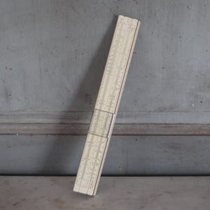 Może przedstawiać: Zabytkowa suwmiarka, instrument matematyczny, jest prezentowana na szarym tle. Suwak jest prostokątny, z kremowym korpusem i czarnymi oznaczeniami. Jest ustawiony pod kątem, prezentując szczegółowe skale i podziały, używane do obliczeń.