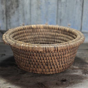 May include: A round, woven basket with a natural, light brown color. The basket has a wide, open top and a slightly flared rim. The weave pattern is visible, creating a textured surface. The basket is likely made of natural fibers.