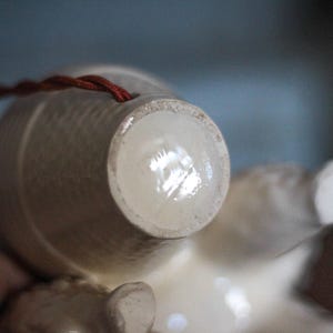 May include: Close-up of a white ceramic figurine, possibly a decorative ornament. The figurine has a textured surface and a small hole with a red string attached. The focus is on the base of the figurine.