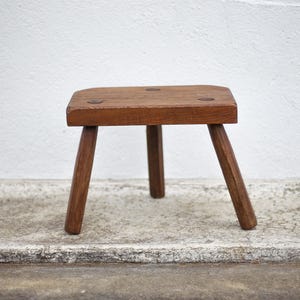 Puede incluir: Un pequeño taburete de madera de tres patas con asiento cuadrado. El taburete es de madera marrón cálido y tiene un aspecto rústico y artesanal. Las patas están anguladas para mayor estabilidad. Este taburete es adecuado para uso decorativo o funcional.