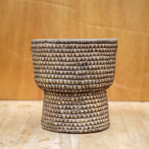 May include: A woven brown basket with a wide, round top and a shorter, cylindrical base. The basket is made of natural materials and has a rustic, handcrafted look.