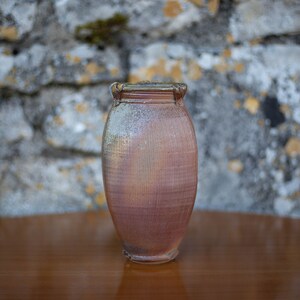 May include: A handmade, textured ceramic vase in reddish-brown. The vase has a slightly flared rim and a rounded body, placed on a wooden surface. The background features a stone wall.