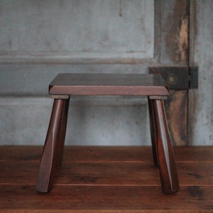 May include: A small, dark brown wooden stool with a square seat and four angled legs. The stool is made of solid wood and has a simple, minimalist design. It is placed on a wooden surface.
