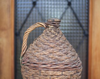 May include: A brown woven wicker bottle with a handle. The bottle is sitting on a dark brown wooden surface.