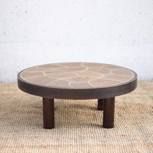 Peut inclure: Une table basse ronde dotée d'un cadre en bois brun foncé et de trois pieds cylindriques. La surface du plateau est incrustée de pierres lisses de couleur beige, disposées en mosaïque avec des joints de couleur claire entre elles.