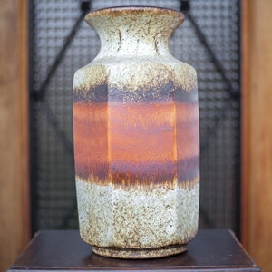 May include: A brown, white, and orange ceramic vase with a geometric shape. The vase is sitting on a dark brown wooden table with a drawer.