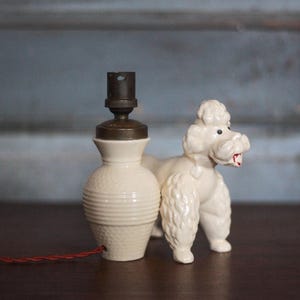 May include: A vintage cream-colored ceramic lamp with a brown top and a red cord. A white poodle figurine stands beside the lamp. The lamp has a textured base and a classic design.