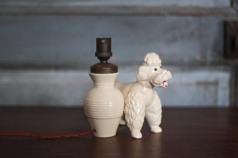 May include: A vintage cream-colored ceramic lamp with a brown top and a red cord. A white poodle figurine stands beside the lamp. The lamp has a textured base and a classic design.